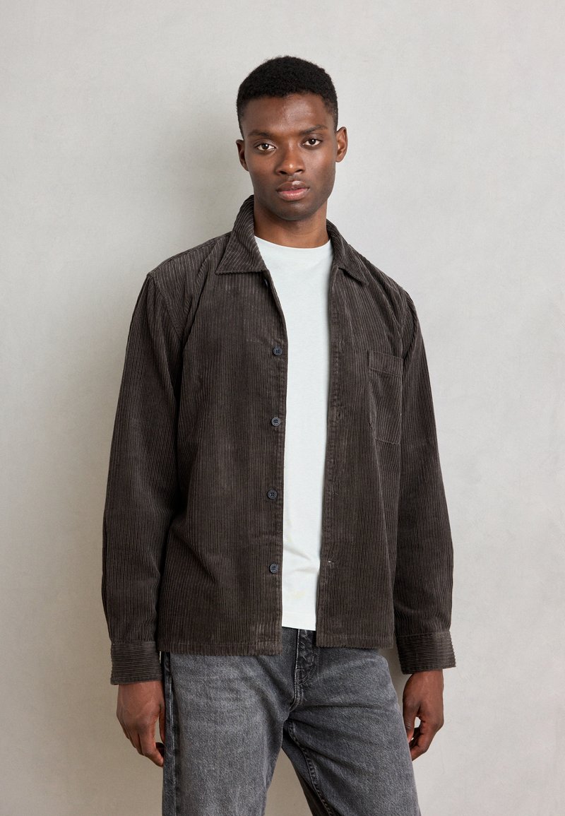 Corduroy shirt in dark brown with button-up front, pointed collar, and chest pocket. Layered over a light grey t-shirt, paired with grey denim.
