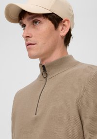 Beiger Strickpullover mit hohem Kragen und Reißverschluss. Waffelstruktur, kombiniert mit einer hellbeigen Mütze. Verfügt über dezente Details.