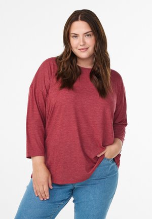 Red, long-sleeve, loose-fitting t-shirt with a soft texture, paired with light blue jeans, model standing with hand on hip.