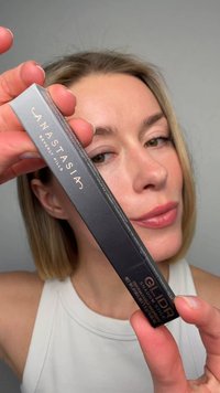 Femme aux cheveux blonds tenant le stick de fard à paupières Anastasia Beverly Hills Glider près de son visage sur un fond gris uni.