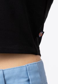 Black cropped t-shirt with a textured finish, featuring a stitched hem. A blue waistband with a smooth finish sits just above the midriff.