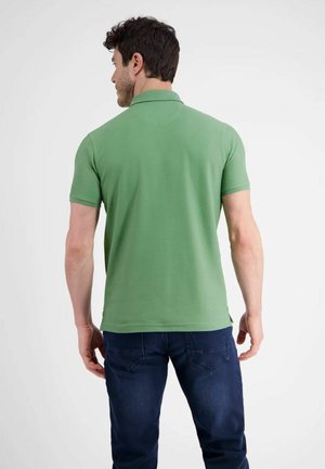 Ein Mann steht mit dem Rücken zum Betrachter, trägt ein grünes Polohemd mit kurzen Ärmeln und dunkelblaue Jeans vor einem schlichten weißen Hintergrund.