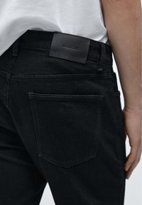 Černé džíny z denimu s hladkou texturou, s koženou etiketu na pasu. Zadní kapsa má klasický prošitý design.