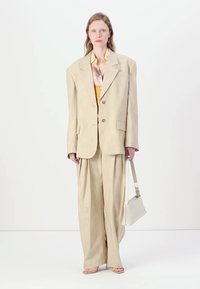 Μπεζ υπερμεγέθη σακάκι με φαρδιές ώμους, συνδυασμένα φαρδιά παντελόνια και κίτρινο τοπ από κάτω. Τα αξεσουάρ περιλαμβάνουν μια λευκή τσάντα.