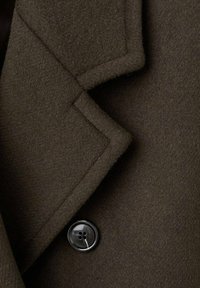 Szczegół ciemnooliwkowego wełnianego płaszcza z wyciętym kołnierzem i czarnym czterodziurkowym guzikiem. Tekstura wydaje się miękka i gęsta.
