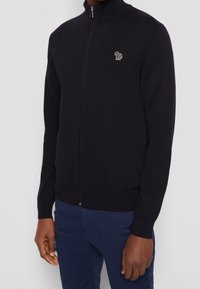 Hombre vistiendo un suéter negro con cremallera completa y un pequeño logo de cebra colorido en el lado izquierdo del pecho, con pantalones azul marino, de pie frente a un fondo gris.