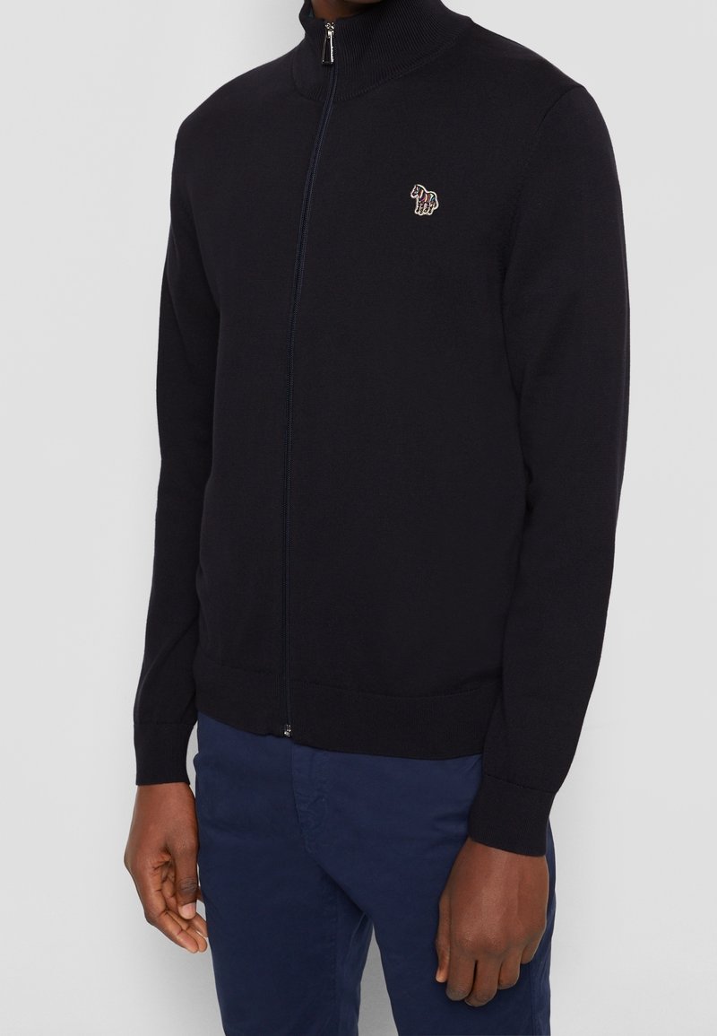 Hombre vistiendo un suéter negro con cremallera completa y un pequeño logo de cebra colorido en el lado izquierdo del pecho, con pantalones azul marino, de pie frente a un fondo gris.