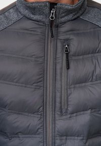 Veste isolée grise avec un col montant, un design matelassé horizontal et des poches zippées. Présente des panneaux et des accents texturés gris sur les épaules.