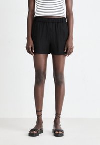 Shorts noirs texturés avec une taille élastique, associés à des sandales noires à lanières. Le sujet porte un haut rayé noir et blanc.