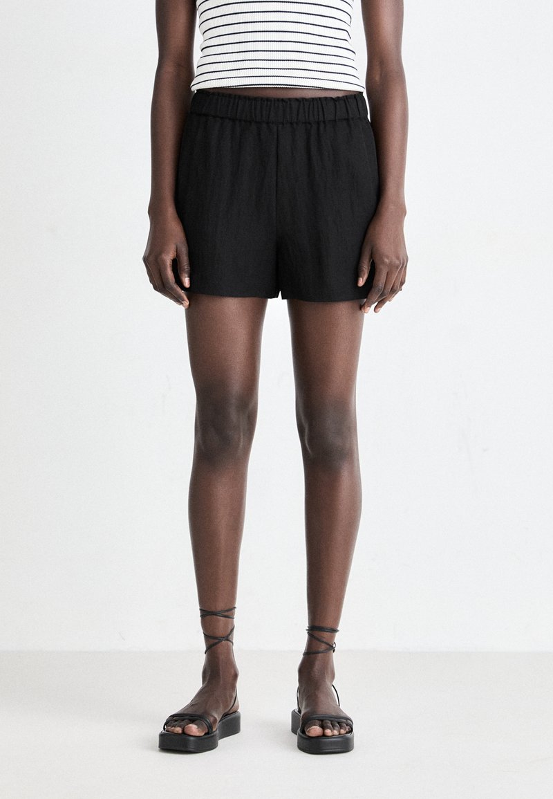Shorts noirs texturés avec une taille élastique, associés à des sandales noires à lanières. Le sujet porte un haut rayé noir et blanc.