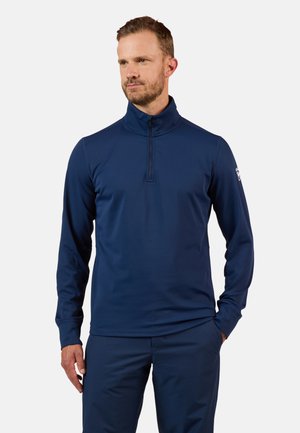 Mann trägt marineblauen Pullover mit Viertelreißverschluss, steht mit einer Hand in der Tasche und schaut leicht zur Seite.