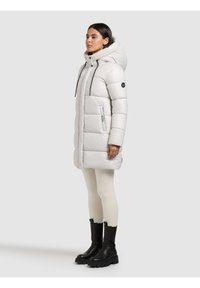 Žena stoji u bijelom puffer kaputu s kapuljačom, kremasti helankama i crnim debelim čizmama na jednobojnoj svijetlosivoj pozadini.