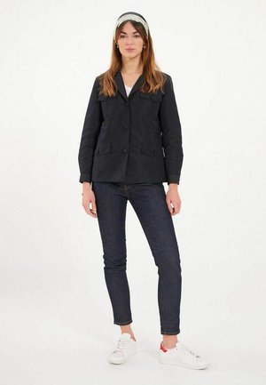 Veste noire avec deux poches avant, fermeture à boutons et design à col, associée à un pantalon en jean sombre et des baskets blanches.