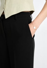 Pantaloni neri su misura con tasche laterali, abbinati a una camicetta verde chiaro cropped. Un braccialetto d'oro con un pendente a forma di cuore adorna il polso.