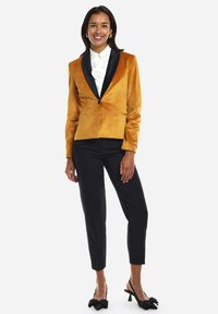 Blazer de terciopelo ámbar con solapas de satén negro, combinado con pantalones ajustados negros y tacones con lazo negro. Camisa blanca de botones debajo.