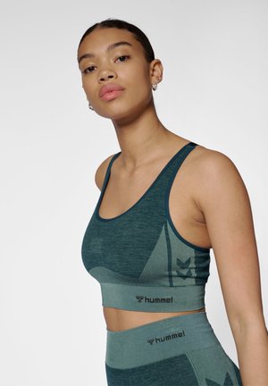 Kobieta ubrana w turkusowy bezszwowy sportowy biustonosz i wysokie legginsy z logo Hummel, pozująca na jednolitym jasnym tle.