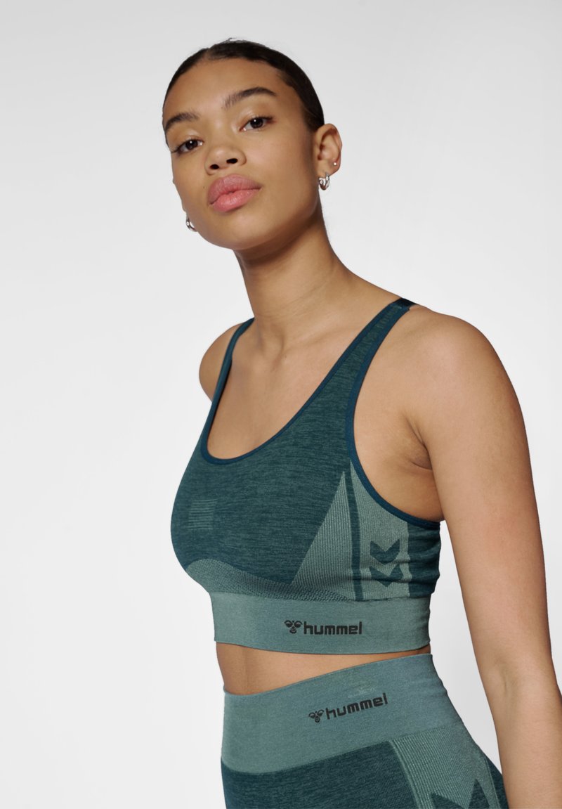 Kobieta ubrana w turkusowy bezszwowy sportowy biustonosz i wysokie legginsy z logo Hummel, pozująca na jednolitym jasnym tle.