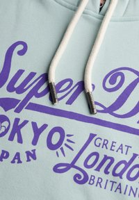 Jasnoniebieska bluza z kapturem z białymi sznurkami, z dużymi fioletowymi grafikami tekstowymi z napisami „Super Dry”, „Tokio” i „Londyn”. Metalowe końcówki na sznurkach.