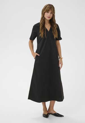 Žena oblečená v čiernych midi šatách s véčkovým výstrihom, krátkym rukávom a bočnými vreckami, so štýlovými čiernymi špicatými balerínkami, stojaca pred jednoduchým pozadím.