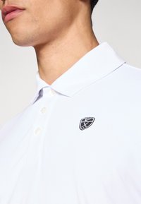 Polo blanc avec un col classique et trois boutons, orné d'un petit logo noir brodé sur la poitrine gauche. Texture de tissu lisse.