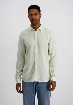 Mann trägt ein langärmliges cremefarbenes Hemd mit Knöpfen und blaue Jeans, steht vor einem einfarbigen hellen Hintergrund.