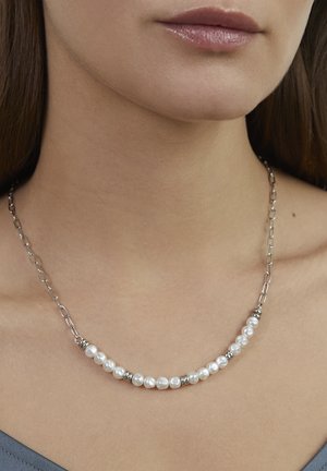 Silberne Halskette mit einer Reihe weißer Perlen und strukturierten Akzenten zwischen den Perlen, getragen an einem hautfarbenen Hals vor einem grauen Oberteil.