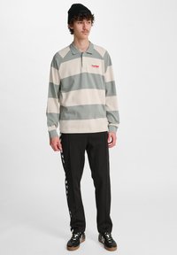 Groen en beige gestreepte polo met lange mouwen, voorzien van een kraag en logo, gecombineerd met zwarte broek met een witte zijkant, en zwarte sneakers.