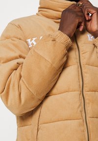 Persona di carnagione scura che sta zipando una giacca imbottita in corduroy beige con polsini elastici e lettere bianche ricamate parzialmente visibili.