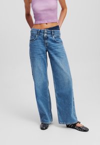 Široké modré džíny vyrobené z denimu, s vysokým pasem, dvěma předními kapsami a zapínáním na knoflík. Spárované s lehkým fialovým tílkem.