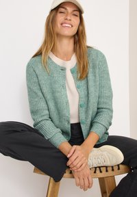 Cardigan en tricot vert clair avec des boutons, tissu texturé, manches longues, porté sur un haut crème, associé à un pantalon foncé et des baskets beiges.