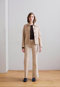 Chaqueta cropped beige con cierre de botones, combinada con pantalones acampanados a juego. Top marrón debajo, diseño elegante, zapatos planos con acentos azules.