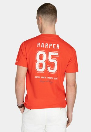 Hombre con camiseta deportiva naranja brillante con "HARPER 85" y "Classic Sports, Timeless Style" impresos en la espalda, combinado con pantalones cortos blancos.