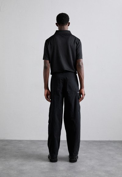 Svart kortermet polo-shirt og svarte cargo-bukser med lommer og avslappet passform. Stoffet ser glatt ut med en matt finish.