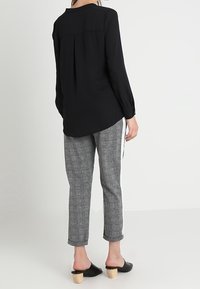 Blusa nera a maniche lunghe con piega sul retro, abbinata a pantaloni grigi a scacchi e mule neri con tacco, che mostrano una vestibilità rilassata.