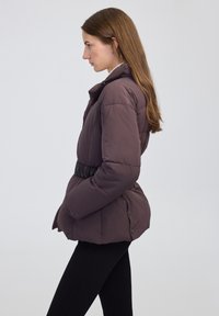 Mujer con cabello largo castaño que lleva una chaqueta acolchada oscura de color malva con cinturón y pantalones negros, girando hacia la izquierda contra un fondo claro y simple.