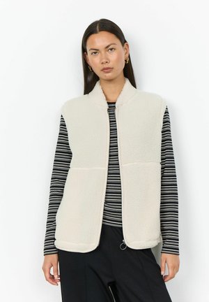 Woman with long dark hair wearing gold hoop earrings, a white fleece vest over a black and white striped long-sleeve top, and black pants.