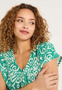 Top de tela con estampado verde, escote en V, mangas cortas y un detalle fruncido en el pecho. Se pueden ver rizos en el cabello y un maquillaje suave.