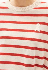 Zbliżenie beżowego swetra z dzianiny z poziomymi czerwonymi paskami oraz małą białą łatą w kształcie wstążki na piersi.