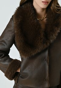 Giacca di pelle marrone con un ampio colletto di pelliccia e polsini abbinati, caratterizzata da una texture liscia e una silhouette rilassata. Dettaglio della collana in catena dorata.