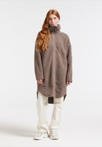 Brun fleece lang frakke med høj krave, frontlynlås og sidelommer. Båret med lyse bukser og sneakers.