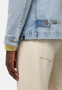 Vaaleansininen farkkutakki, jossa on napillinen läppä, kontrastiset keltaiset hihansuut ja näkyvät ompelusaumat. Yhdistetty vaaleanbeigeihin housuihin.