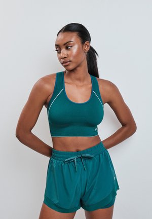 Femme athlétique aux cheveux foncés en queue de cheval portant un soutien-gorge de sport bleu sarcelle et un short assorti, debout les mains derrière le dos, regardant vers la gauche.