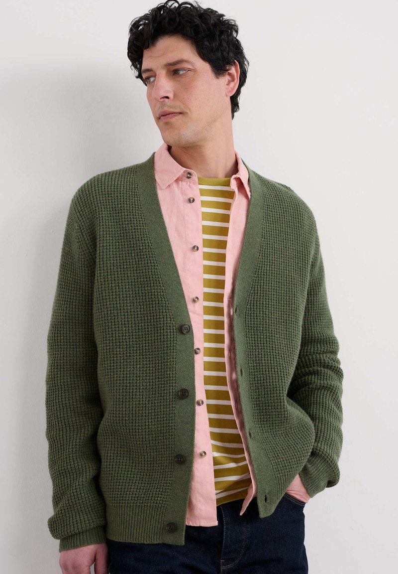 Cardigan tricotat verde, cu decolteu în V, închidere cu nasturi și manșete cu nervuri, purtat peste o cămașă roz cu nasturi și un tricou în dungi galben-verde.