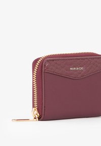 Portafoglio color bordeaux con dettaglio in pelle testurizzata, cerniera dorata e piccolo logo. Forma rettangolare compatta con finitura lucida.