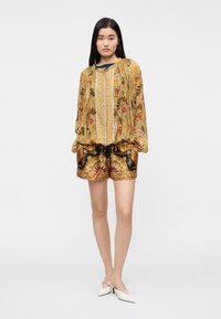 Αέρινη κίτρινη μπλούζα με λουλουδάτα σχέδια, σε συνδυασμό με καφέ σορτς με σχέδια. Λευκά τακούνια ολοκληρώνουν το σύνολο, παρουσιάζοντας ένα μείγμα χρωμάτων και υφών.