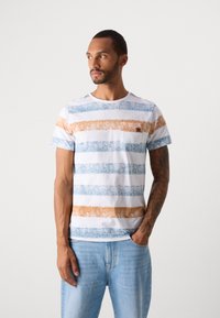 Mann med tatovert arm iført hvit T-skjorte med blå og oransje striper og lyseblå jeans, ser til venstre med hånden i lomma.