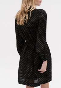 Zwarte jurk met gouden stippen, lange mouwen en een cinch taille. Heeft een knoopsluiting bij de manchetten en een vloeiende, doorschijnende textuur aan de zoom.