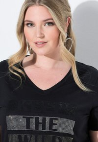 Černé tričko s výstřihem do V a zdobeným textem, který obsahuje směs stříbrných a černých krystalů. Dlouhé vlasy a náušnice ve tvaru srdce jsou viditelné.