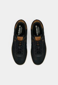 Ζευγάρι μαύρα αθλητικά παπούτσια Adidas με καφέ δερμάτινες λεπτομέρειες, μαύρα κορδόνια και σόλες από καουτσούκ, σε λευκό φόντο, φωτογραφημένα από πάνω.