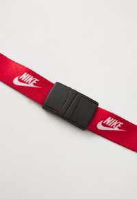 Červený nylonový pásek s černou plastovou sponou, s opakujícími se bílými logy Nike podél pásku. Plackový design a texturovaný povrch.